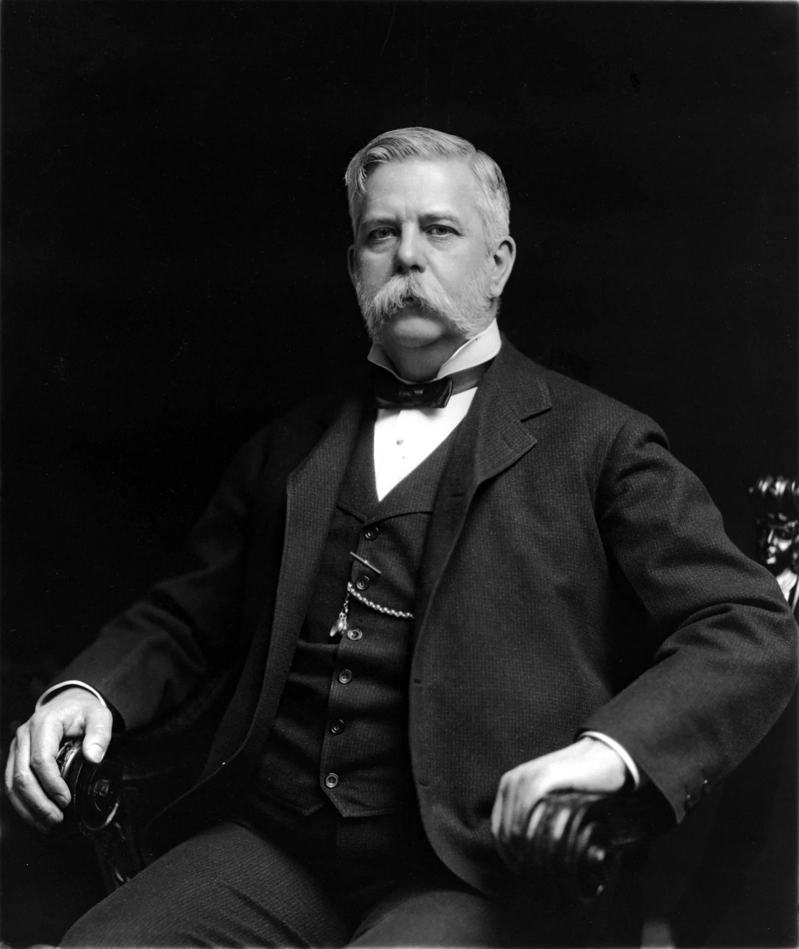 Studio photograph of a seated middle-aged man against a black background. He has white hair brushed back, a heavy white moustache and side-whiskers, and wears a dark three-piece suit with a white shirt, a dark bow tie and a watch chain across the waistcoat. Both hands rest on the arms of a carved wooden chair. He looks directly at the camera and is not smiling.