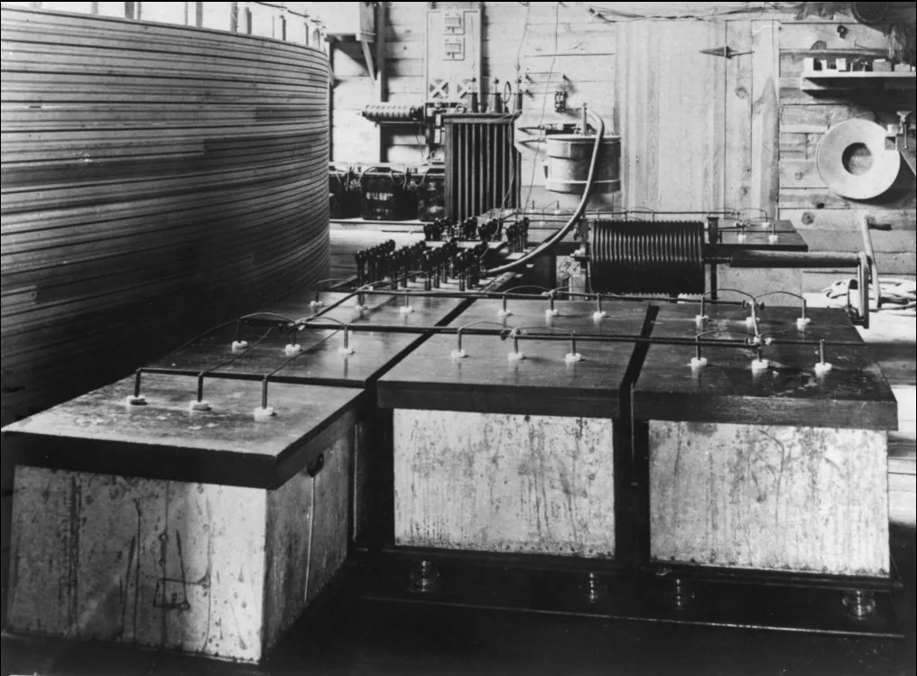 Interior of a timber building. In the foreground three large pale rectangular tanks stand side by side on a dark base; bare metal rods run between ceramic standoffs across their tops. Behind them, a finned transformer, a metal bucket, a dark cylindrical coil and a switch panel stand against a plank wall. Filling the whole left edge is the curved wooden wall of a very large coil, its horizontal turns running out of frame.