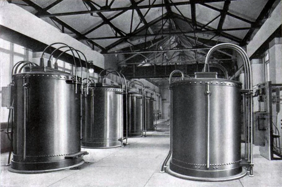 Interior of a brick hall with a light steel roof truss. Six tall riveted steel cylinders, each about the height of a person, stand in two rows down the room on low plinths. Heavy insulated conductors curve up out of the top of each one and run across to switch panels on the walls. Daylight comes from windows at the left; there is nobody in the room.