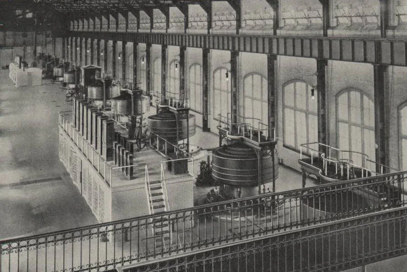 View down the length of a tall generator hall, taken from a gallery above. A row of large circular machine housings, each the height of a person and ringed with vertical ribs, runs away into the distance along a raised floor with a white-painted railing. Tall arched windows line the far wall. A short flight of steps leads up to the floor; one figure stands between two of the machines.