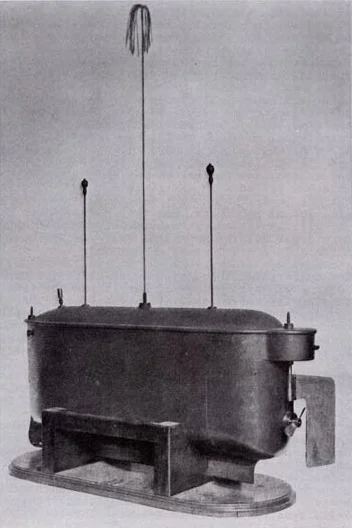 Photograph of a small boat-shaped metal hull with a flat lid, standing on a low wooden cradle on a plain pale background. Three slender vertical rods rise from the lid, the centre one taller than the other two, each ending in a small knob. A flat rudder blade projects from the stern, and a knurled fitting sits on the deck beside it. Nothing is connected to the hull.
