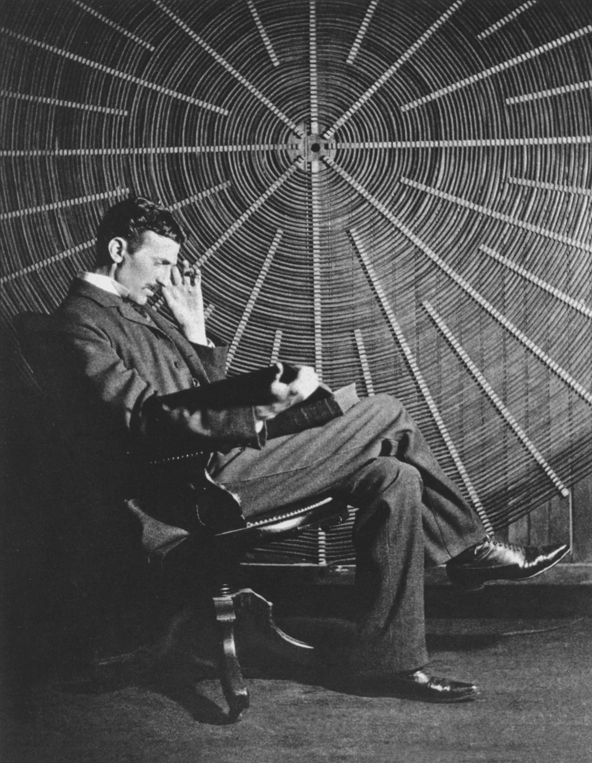 Photograph of a man in a dark suit sitting sideways in a small wooden chair, legs crossed, reading an open book held in his lap with one hand raised to his temple. Filling the whole wall behind him is an enormous flat spiral of wire, many turns wide, with straight spokes radiating from its centre like the ribs of a wheel.